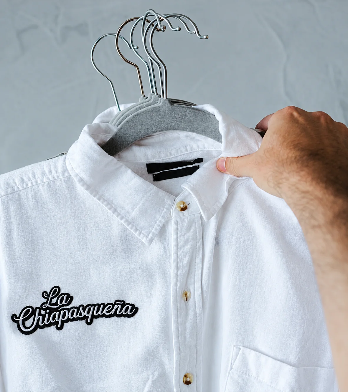 Camisa blanca con el logotipo de La Chiapasqueña bordado, colgada en un gancho.