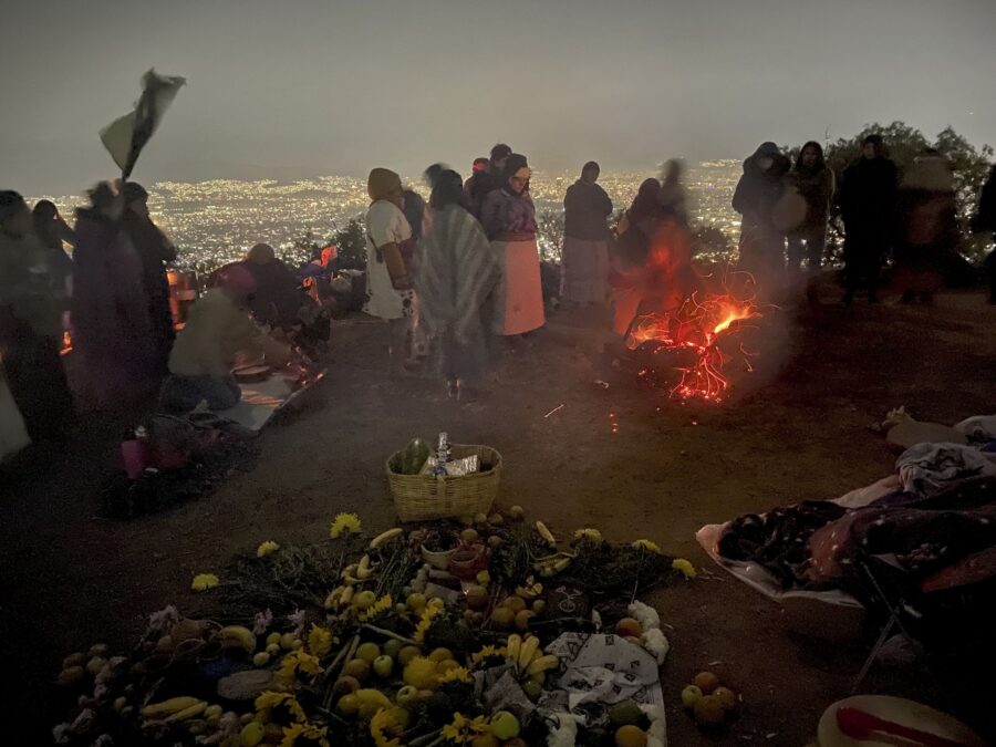Personas reunidas de madrugada alrededor de una ofrenda de flores y una fogata, con la ciudad iluminada al fondo